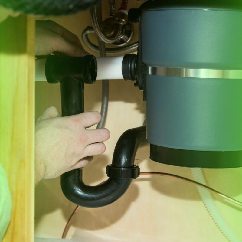 A technician adjusting the black PVC drain pipe under a garbage disposal unit beneath a kitchen sink.