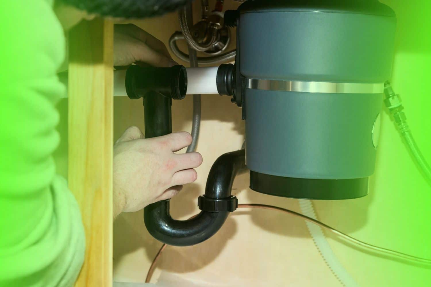 A technician adjusting the black PVC drain pipe under a garbage disposal unit beneath a kitchen sink.