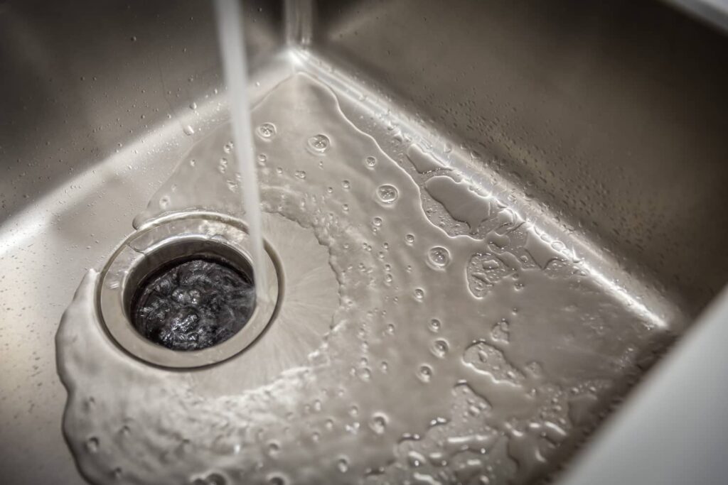Water running from a faucet into a stainless steel kitchen sink and flowing down a garbage disposal drain.