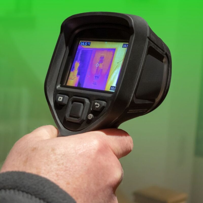A hand holding a handheld thermal imaging camera displaying an infrared heat map scan of an interior doorway.