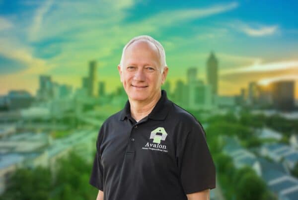 A smiling male home inspector wearing a black polo shirt with the Avalon Home Inspections Inc. logo, set against a blurred city skyline background.