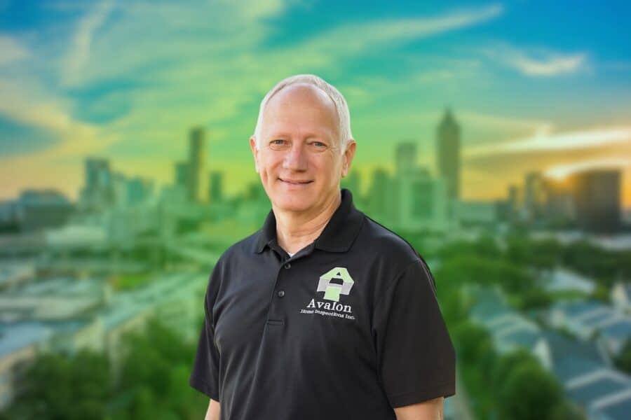 A smiling male home inspector wearing a black polo shirt with the Avalon Home Inspections Inc. logo, set against a blurred city skyline background.