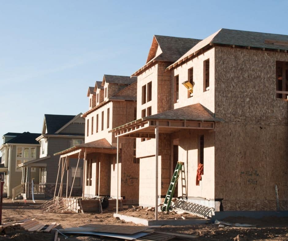 Row of newly framed two-story houses under construction with exposed plywood sheathing, unfinished window openings, and ladders on a dirt lot.
