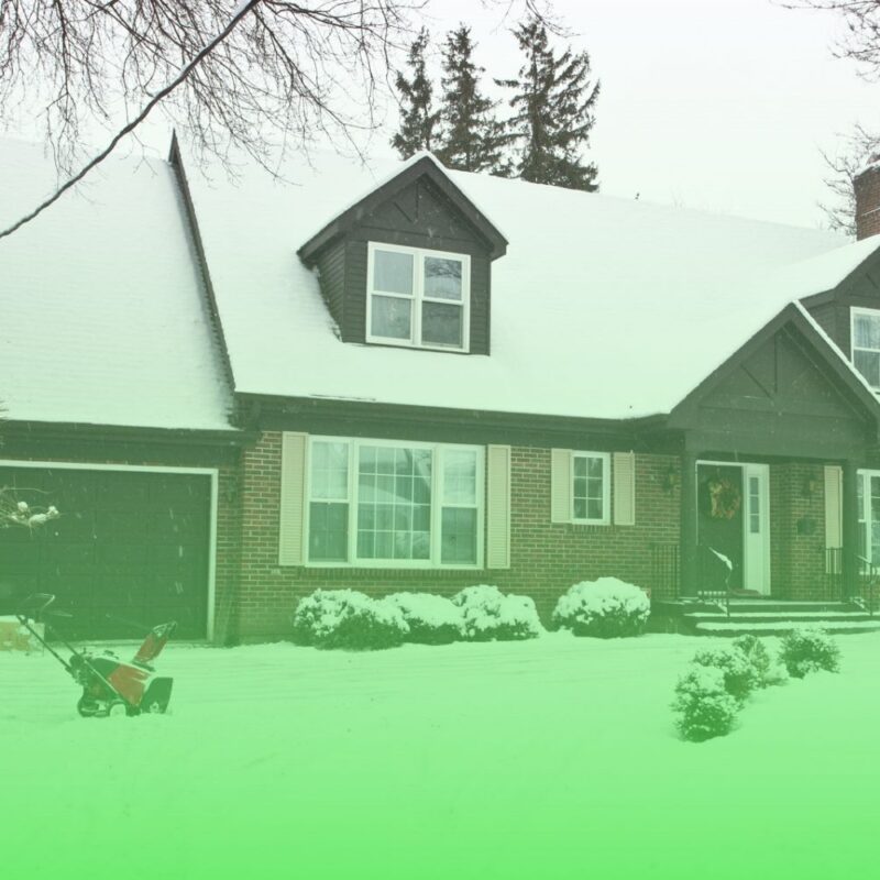 Two-story brick home covered in fresh winter snow with a snowblower parked in the snowy front yard and driveway.